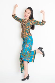 Portrait of an Southeast Asian girl in batik dress arms up happy jumping around, full length standing isolated on plain background.