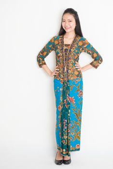 Full body Southeast Asian woman in batik dress standing on plain background.