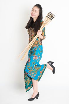 Full body portrait of Southeast Asian woman in batik dress hand holding pelita or oil lamp standing on plain background.