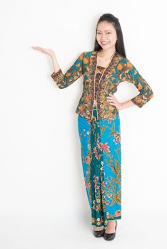Full body portrait of Southeast Asian female in batik dress hand holding something standing on plain background.
