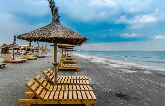 Cloudy seaside beach with wooden lounge chairs and straw umbrellas.