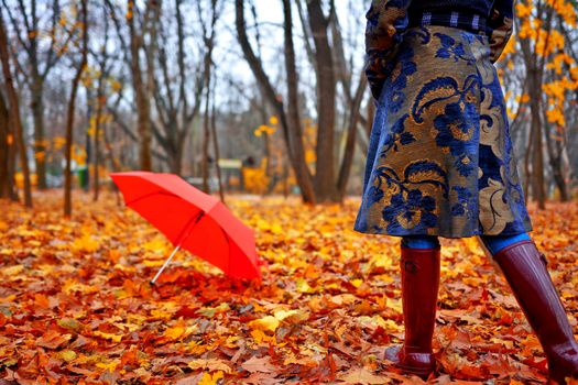 Woman with rubber boots and autumn coat walking over withered yellow leaves towards umbrella .