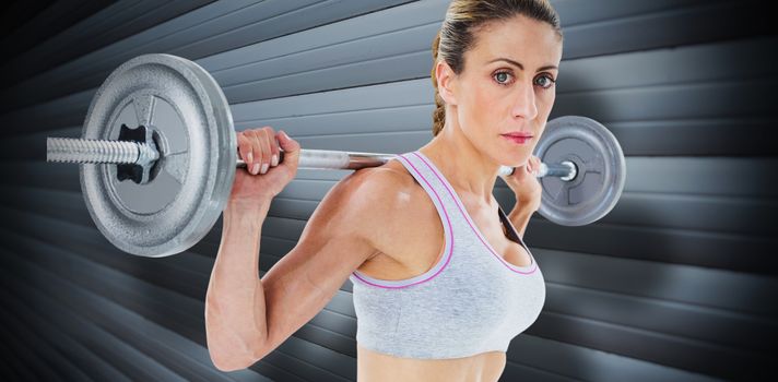 Strong female crossfitter lifting barbell behind head looking at camera against grey shutters