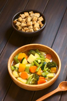 Wooden bowl of vegetable soup made of zucchini, green bean, carrot, broccoli, potato and pumpkin with a small bowl of croutons in the back, photographed on dark wood with natural light (Selective Focus, Focus one third into the soup)