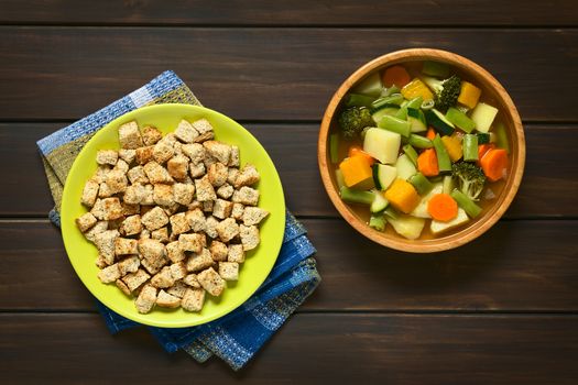 Overhead shot of a plate of homemade croutons and a wooden bowl of vegetable soup made of zucchini, green bean, carrot, potato, pumpkin and broccoli, photographed on dark wood with natural light