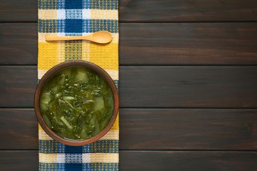 Overhead shot of a rustic bowl of chard soup and a small wooden spoon on kitchen towel, photographed on dark wood with natural light