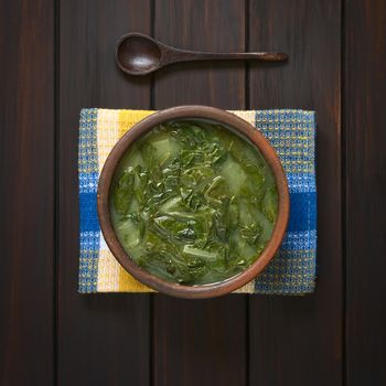 Overhead shot of a rustic bowl of chard soup on kitchen towel with a small wooden spoon above, photographed on dark wood with natural light