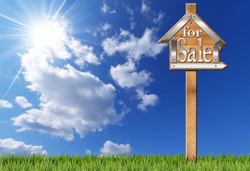 Wooden and metallic sign in the shape of house with text for sale and wooden pole. For sale real estate sign on blue sky with clouds, sun rays and green grass