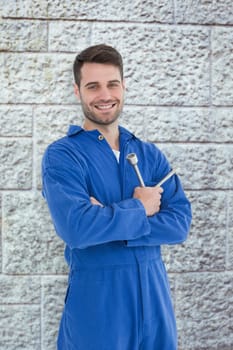 Smiling young male mechanic holding spanner against grey brick wall