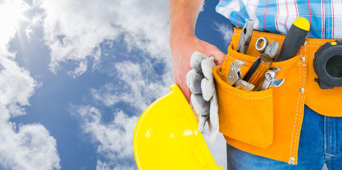 Manual worker wearing tool belt while holding gloves and helmet against bright blue sky with clouds