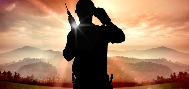 Confident handyman holding drill machine against sun shining over mountains