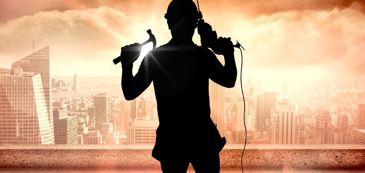Smiling handyman holding hammer and drill machine against sun shining over city