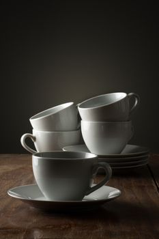 Five plain white ceramic coffee or tea cups on a wooden kitchen counter or table side lit with dramatic lighting with a single cup and saucer in the foreground