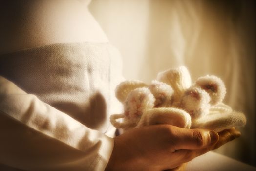 Close up of pregnant woman who is holding baby shoes in front of her baby bump.