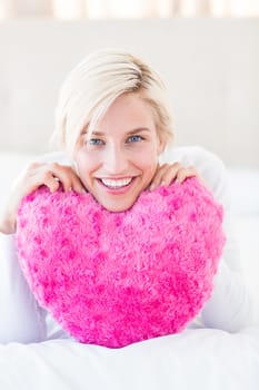 Smiling blonde woman holding heart pillow in the bedroom