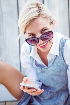 Pretty blonde woman wearing sun glasses and texting with her mobile phone on wooden background