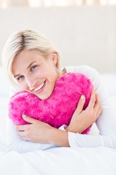 Smiling blonde woman holding heart pillow in the bedroom