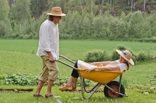 Senior man carries a senior woman in a wheelbarrow outdoors near a vegetable patch. They're laughing and having fun.