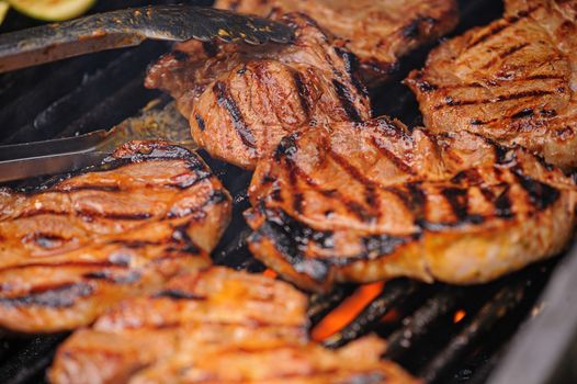 close up of meat grilling on a barbeque. Pair of tongs lifts a steak. Flames are visible under the meat. Narrow depth of field