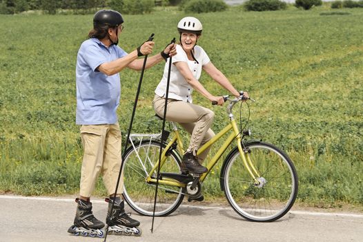 Senior couple vigorously exercising. The man is Nordic inline skating, and the woman is cycling. They're on a country road through green fields.