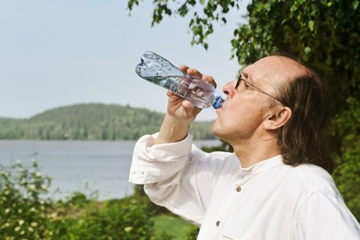 Thirsty senior man drinks water from a bottle outdoors. There is a lake and forest in the background. Digital filter has been used