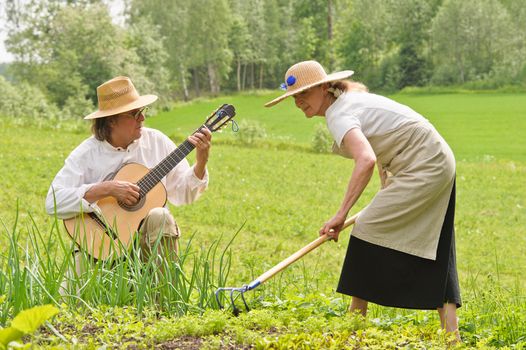 Senior man and woman in a vegetable garden. The woman is raking the soil. The man is playing a guitar