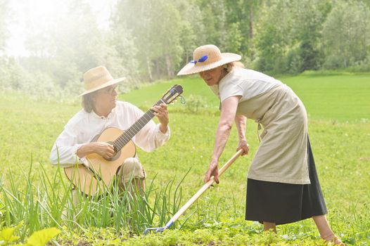 Senior man and woman in a vegetable garden. The woman is raking the soil. The man is playing a guitar. Flare from the top left corner
