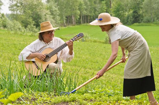 Senior man and woman in a vegetable garden. The woman is raking the soil. The man is playing a guitar