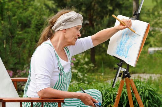 Senior woman painting outdoors. She sits in a canvas chair in front of the easel. The day is overcast giving a soft light