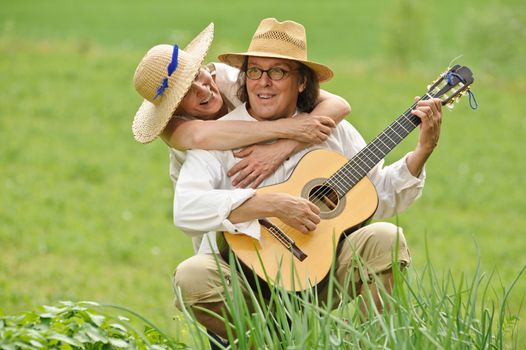 Senior couple having fun together. The man is sitting and playing a guitar. The woman hugs him from behind. They are outdoors.