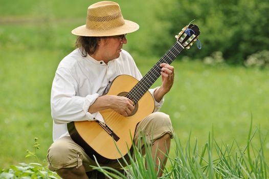 Senior man plays a nylon string guitar outdoors, He is sitting on a log. There's a blurred field in the background