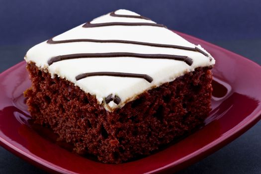 Red velvet cake with white icing and drizzles of chocolate placed on square, red plate on black background.