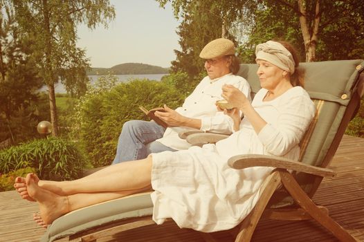 Senior couple relax in deckchairs on a wooden terrace, She is eating from a bowl and he is reading a book. Digital filters used to create a retro look. Lake and forest in the background.