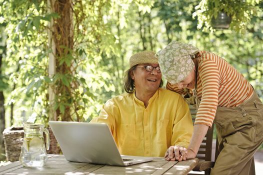 Senior couple outdoors with a laptop, They're laughing. There's a sunny background of trees and bushes