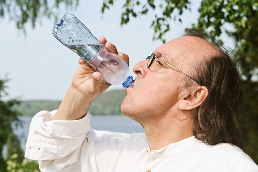 Thirsty senior man drinks water from a bottle outdoors. There is a lake and forest in the background. Digital filter has been used