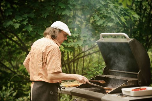 Senior man grilling outdoors. Smoke rises from the barbeque. The picture is desaturated and has been treated with digital filters