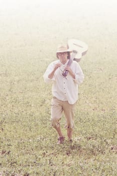 Senior man walks through a field with a guitar across his shoulder. He chews a stalk of grass. Digital filters used to create a faded retro atmosphere.