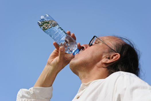 Thirsty senior man drinks water from a bottle outdoors. There is a lake and forest in the background. Low camera angle.