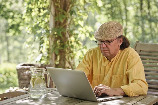 Senior man sits outdoors at a wooden table under a pergola and works on a laptop computer. It's summer and there's a background of green trees and bushes