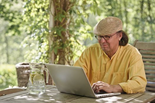 Senior man sits outdoors at a wooden table under a pergola and works on a laptop computer. It's summer and there's a background of green trees and bushes