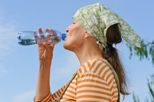 Senior woman drinks water from a bottle outdoors. Blue sky in the background. Low camera angle