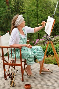 Senior woman painting outdoors. She sits in a canvas chair in front of the easel. The day is overcast giving a soft light