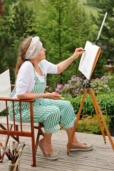 Senior woman painting outdoors. She sits in a canvas chair in front of the easel. The day is overcast giving a soft light