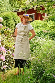 Senior woman standing in her garden, She is surrounded by bushes. There's a red house partly seen in the background. She's wearing an apron