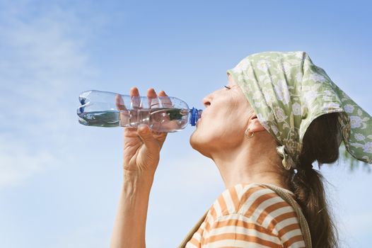 Senior woman drinks water from a bottle outdoors. Blue sky in the background. Low camera angle