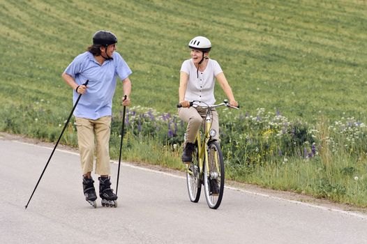 Senior couple vigorously exercising. The man is Nordic inline skating, and the woman is cycling. They're on a country road through green fields.