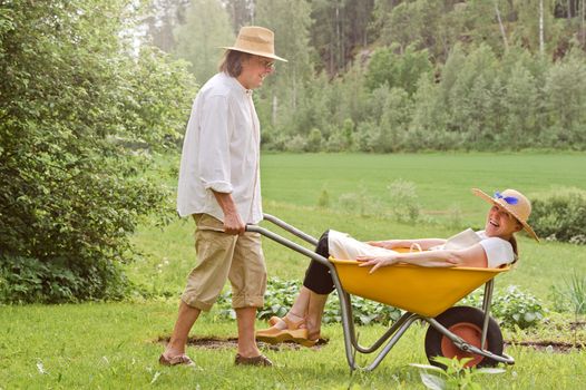 Senior man carries a senior woman in a wheelbarrow outdoors near a vegetable patch. They're laughing and having fun. Digital filtering used