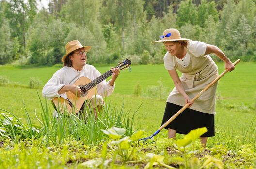 Senior man and woman in a vegetable garden. The woman is raking the soil. The man is playing a guitar