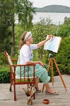 Senior woman painting outdoors. She sits in a canvas chair in front of the easel. The day is overcast giving a soft light