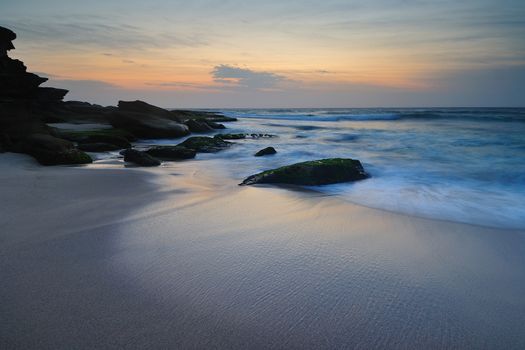 Sunrise seascape with a lone mossy rock with a flowing wave around it